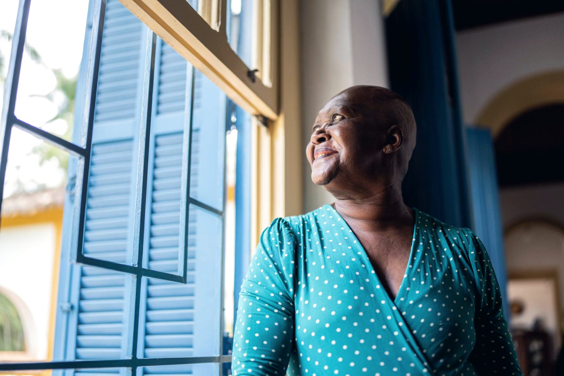 An older woman looks out through blue-shuttered windows.